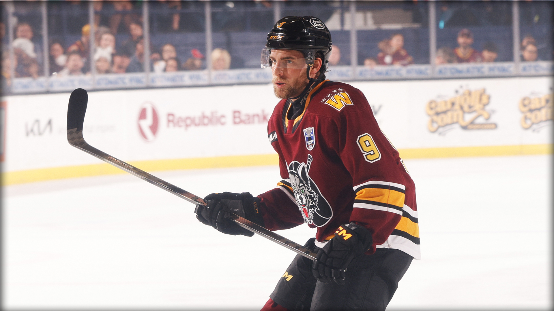 A hockey player in a maroon uniform with the number 9 stands on the ice holding a hockey stick during a game.