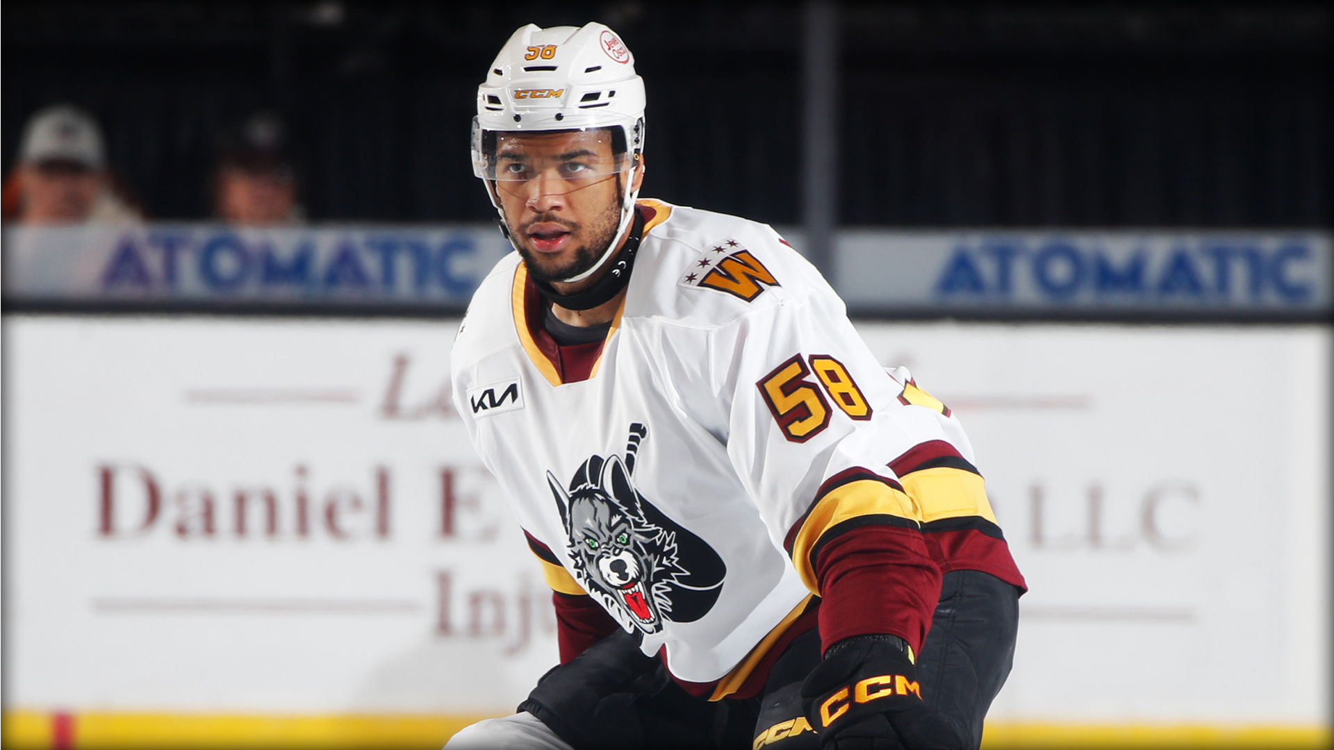 Ice hockey player in white jersey with maroon and yellow trim, number 58, on the ice during a game, wearing helmet and gloves. An advertisement banner is visible in the background.