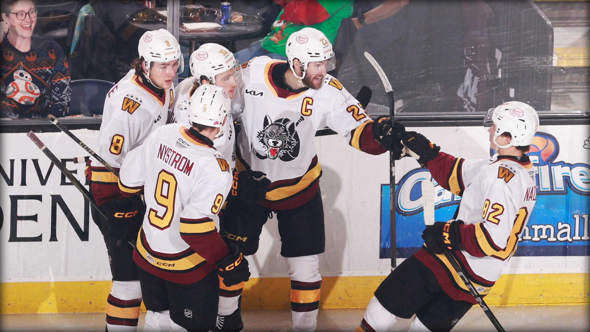 Five ice hockey players in white and maroon jerseys celebrate on the ice near the rink boards while spectators watch from the stands.