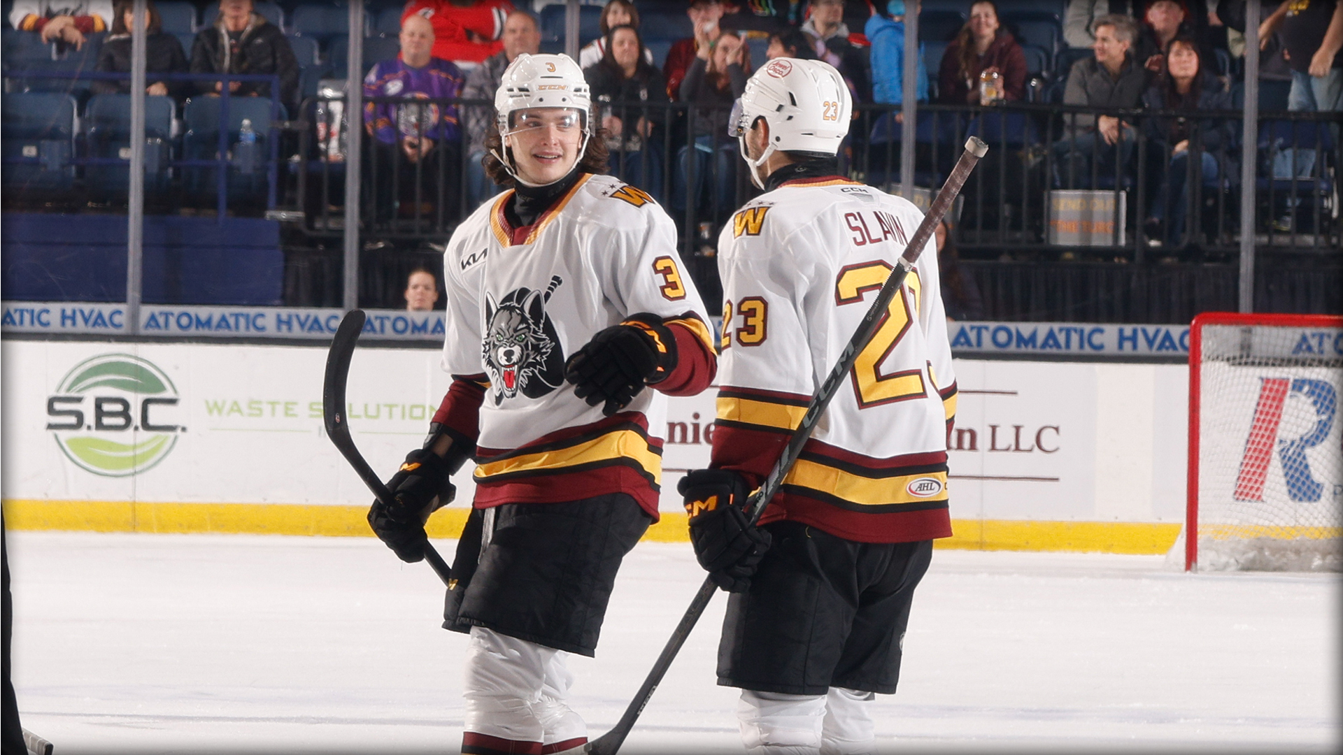 Two ice hockey players in white jerseys stand on the rink, conversing and holding their sticks, with spectators seated in the background.