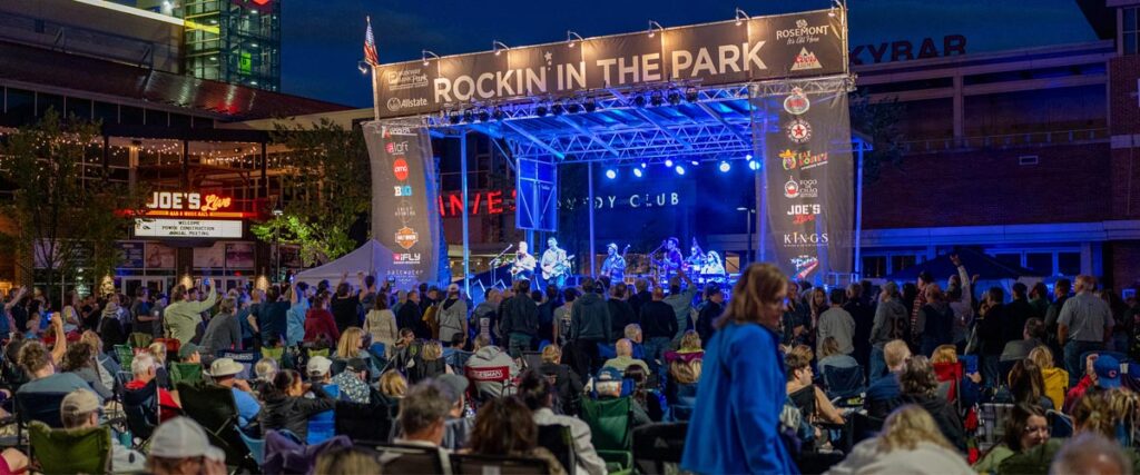 A large crowd gathers outdoors at night in front of a brightly lit stage with a band performing under a banner that reads Rockin in the Park. Buildings and restaurant signs are visible in the background.