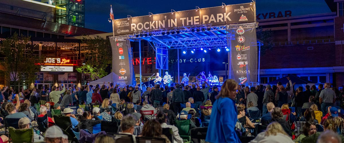 A large crowd gathers outdoors at night in front of a brightly lit stage with a band performing under a banner that reads Rockin in the Park. Buildings and restaurant signs are visible in the background.