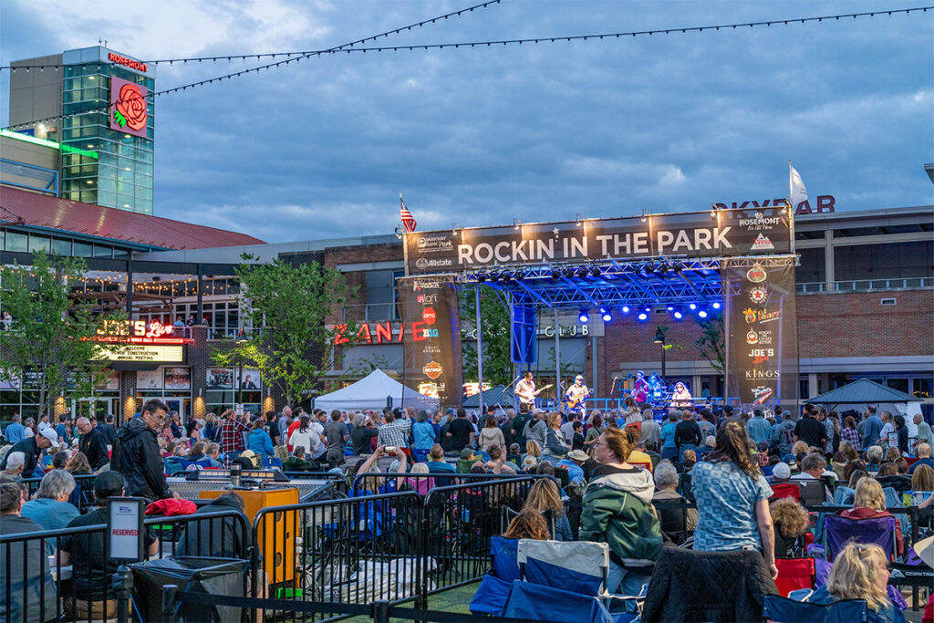 A large crowd gathers at an outdoor concert called Rockin’ in the Park. The stage is lit up with musicians performing, and people are seated and standing under a cloudy evening sky, surrounded by buildings and string lights.