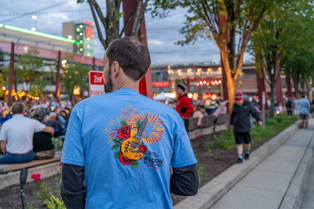 A man in a blue Rockin on the River t-shirt stands near a crowd at an outdoor event with trees, benches, and festive lights in the background. The scene appears lively and relaxed during dusk.