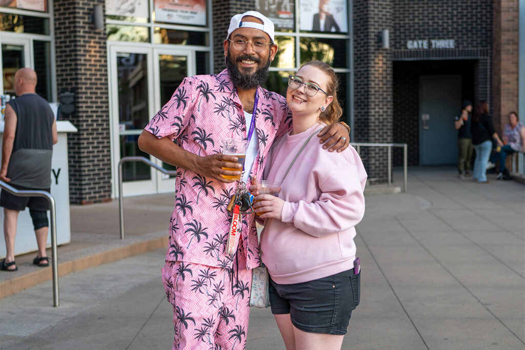 A smiling man in a pink palm tree outfit and a woman in a light pink hoodie stand together outdoors, each holding a drink. The background shows other people, a brick building, and doors marked Gate Three.