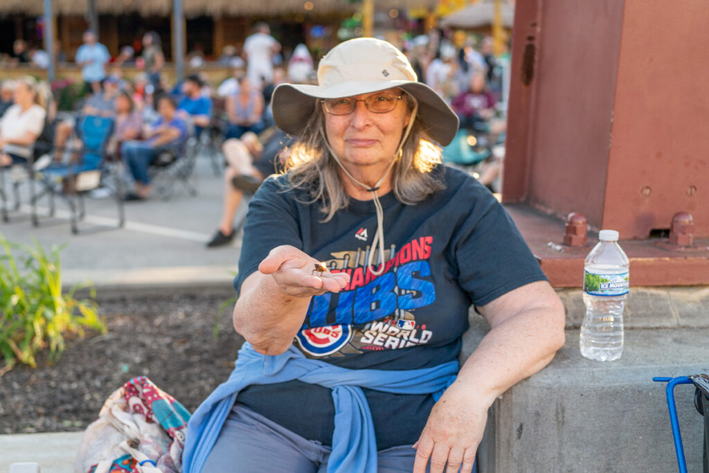 An older woman wearing a sun hat and sunglasses sits outdoors at an event, holding out a coin. She wears a Cubs World Series Champions t-shirt, with a water bottle and bag beside her. People and chairs are in the background.