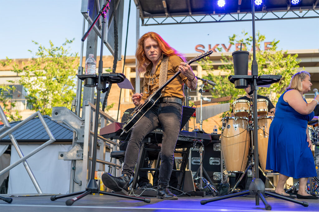 A musician with long red hair plays an electric guitar on an outdoor stage, looking toward the camera. Behind him are drums, keyboards, and another performer in a blue dress. The stage is lit with blue and purple lights.