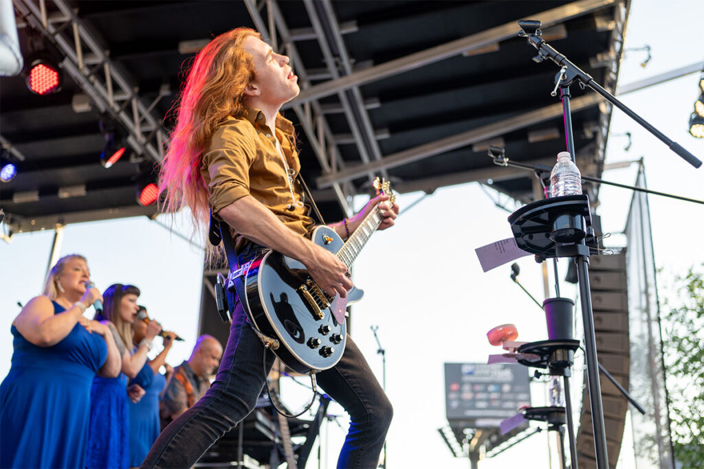 A guitarist with long red hair passionately plays on an outdoor stage, while three backup singers in blue dresses perform in the background. Stage lights and audio equipment are also visible.