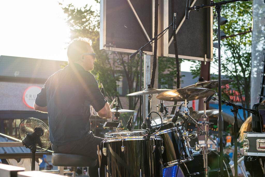 A drummer plays a drum set on an outdoor stage during sunset, with sunlight streaming through trees and a crowd visible in the background.