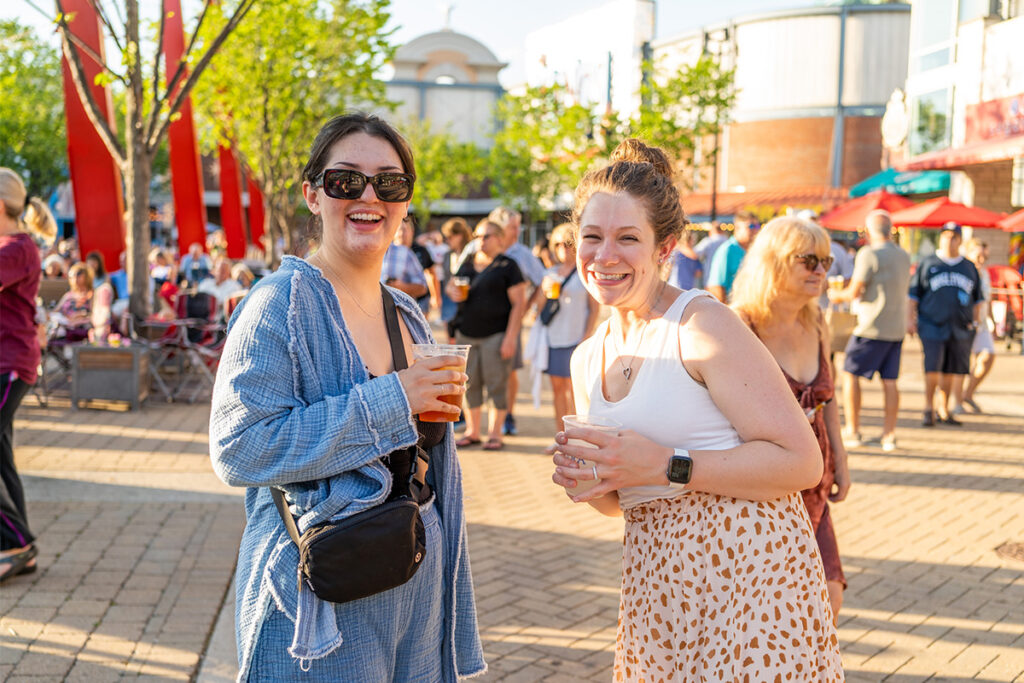 Two women smiling and holding drinks at an outdoor event on a sunny day, surrounded by people and trees in a lively, urban plaza.