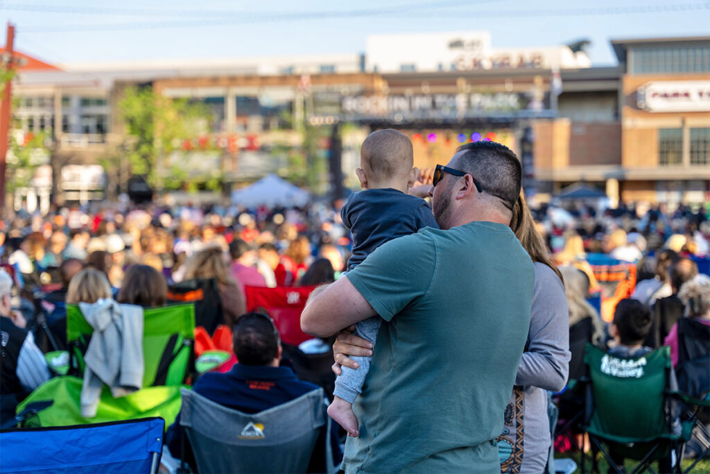 A man holding a baby stands among a crowd seated on colorful folding chairs at an outdoor event, with buildings and a stage visible in the background.