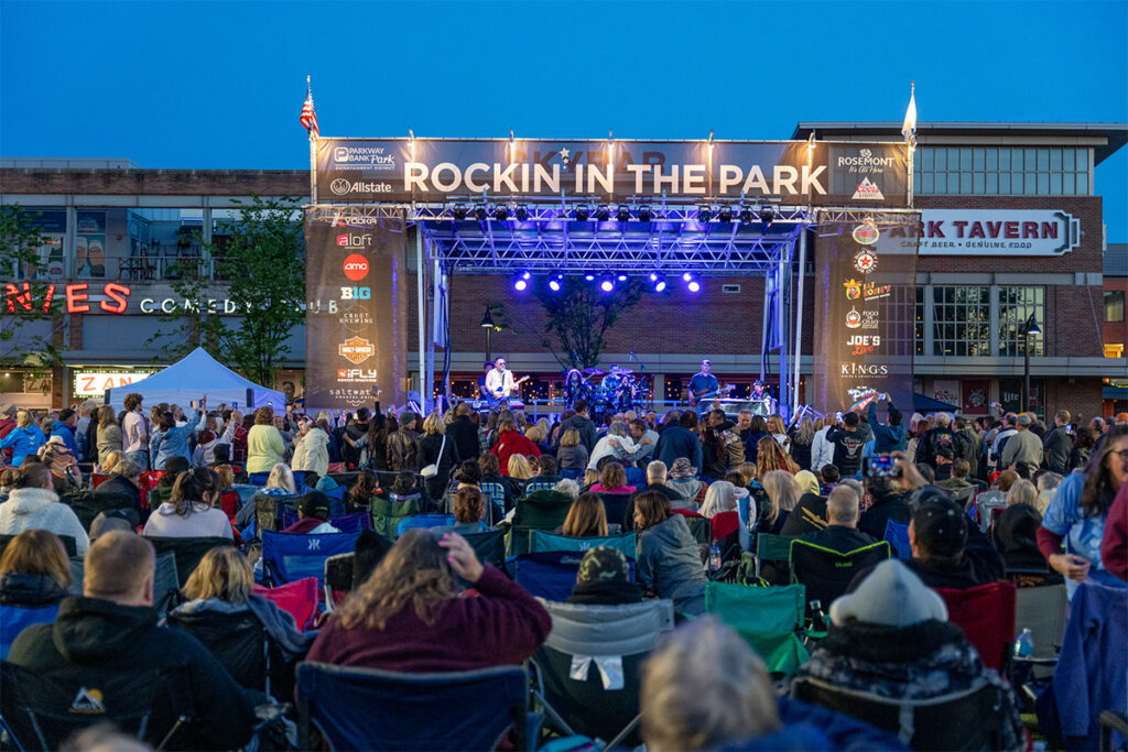 A large crowd gathers in front of a stage with “Rockin’ in the Park” on the banner, watching a live music performance at dusk in an outdoor plaza surrounded by shops and restaurants.