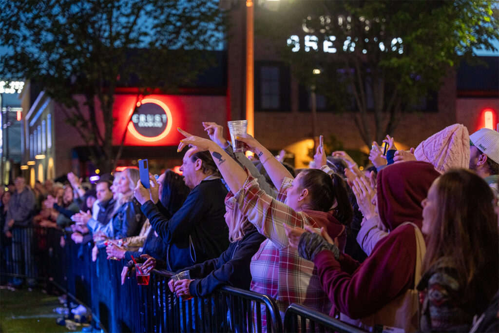 A crowd of people at an outdoor concert or event stands behind a barricade, many holding up phones and drinks, with colorful lights and buildings in the background at dusk.