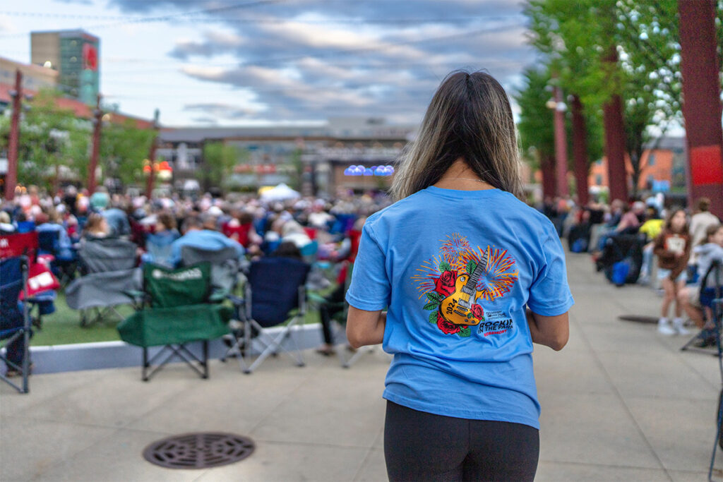 A woman with long hair in a blue T-shirt with a colorful guitar design stands facing an outdoor crowd seated on folding chairs at a music event or festival under a cloudy sky.