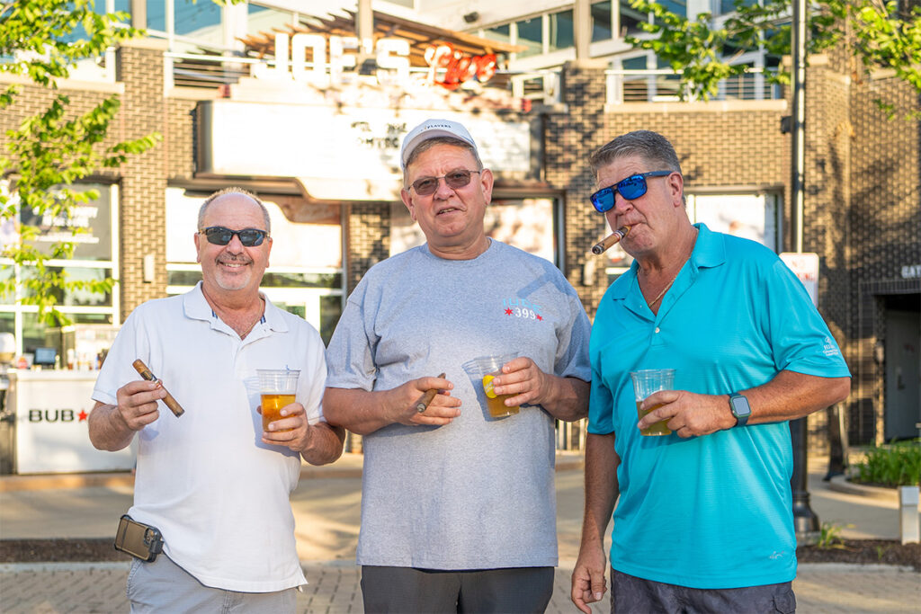 Three older men stand outside, each holding a drink and a cigar, smiling at the camera. They wear casual clothes and sunglasses. A restaurant sign reading JOES is visible in the background.