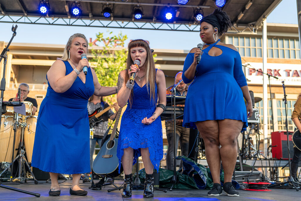 Three women in blue dresses sing on an outdoor stage, each holding a microphone. Musicians and instruments are visible behind them, with a building and trees in the background. Blue stage lights shine above.