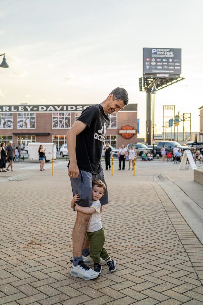 A man wearing a black Adidas shirt stands outdoors, smiling down at a small child who hugs his leg. People, cars, and a Harley-Davidson building are visible in the background on a sunny day.