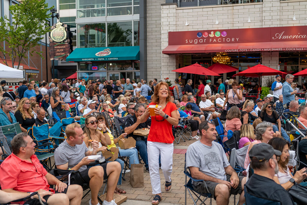 A large crowd gathers outside the Sugar Factory restaurant. People sit in lawn chairs and talk, while a woman in red pants and a white top stands smiling in the center, holding a drink.