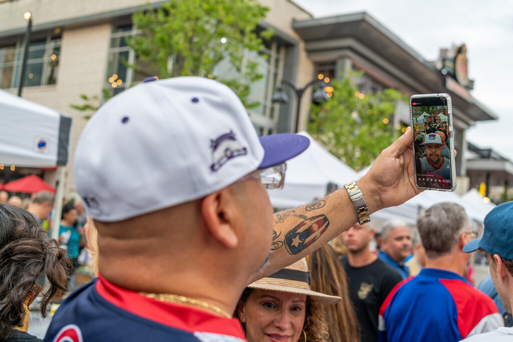 A man wearing a baseball cap and jersey takes a selfie with his phone at an outdoor event, surrounded by a crowd and tents, with buildings visible in the background.