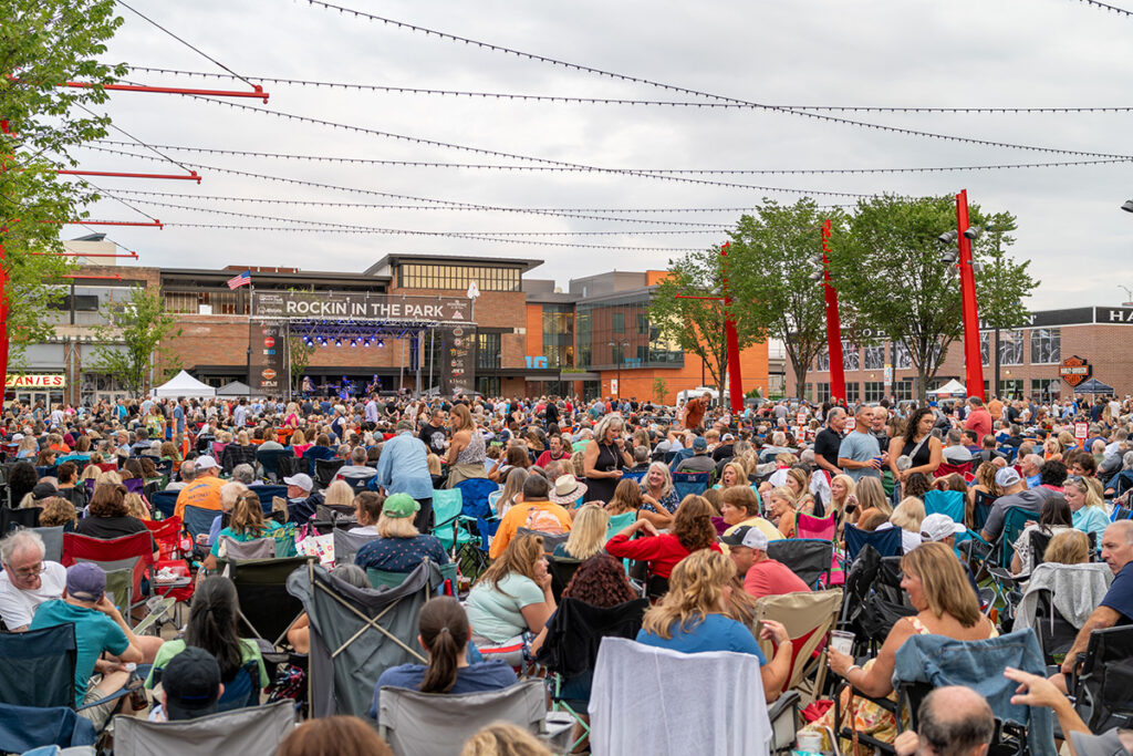 A large crowd sits on lawn chairs facing a stage at an outdoor concert called Rockin’ in the Park, with people chatting and enjoying the event under a cloudy sky. Buildings and string lights surround the plaza.