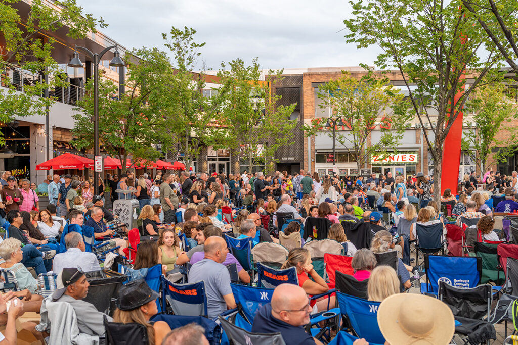 A large crowd of people sits outdoors in lawn chairs at an event in a city plaza, surrounded by trees and shops under a cloudy sky. Many people are talking and enjoying the atmosphere.
