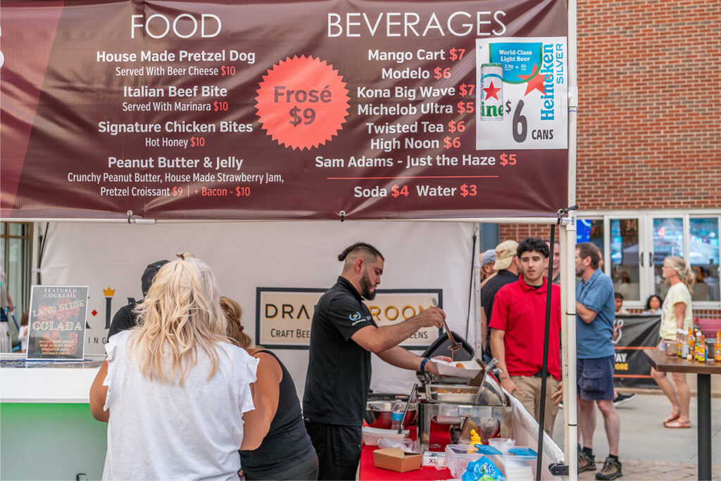 A man prepares food at an outdoor vendor stall with a menu above listing food and beverages. Several people wait nearby, and brick buildings are visible in the background.
