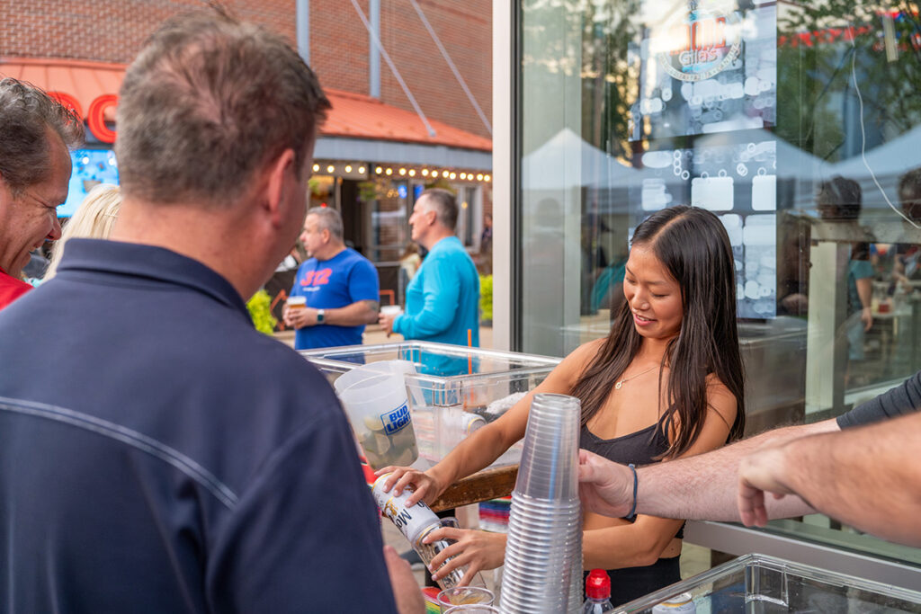A woman behind a counter pours a drink from a can while smiling at an outdoor event. Several people stand in front of her, and clear plastic cups are stacked on the counter. Tents and buildings are visible in the background.