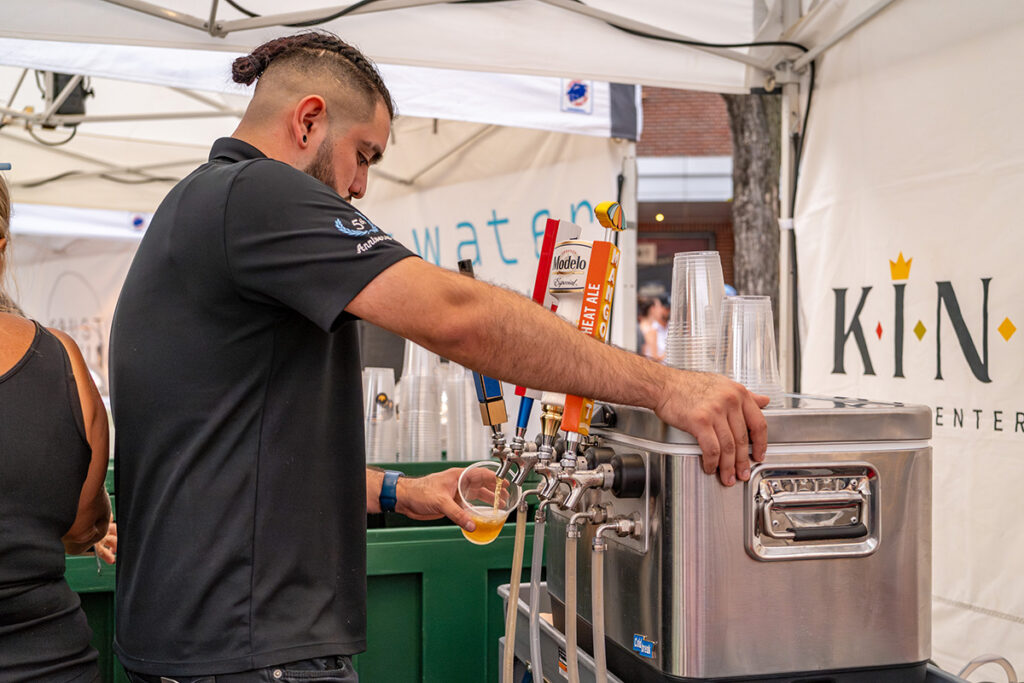 A man in a black polo shirt pours beer from a tap into a plastic cup at an outdoor event booth with plastic cups stacked beside him and branded banners in the background.