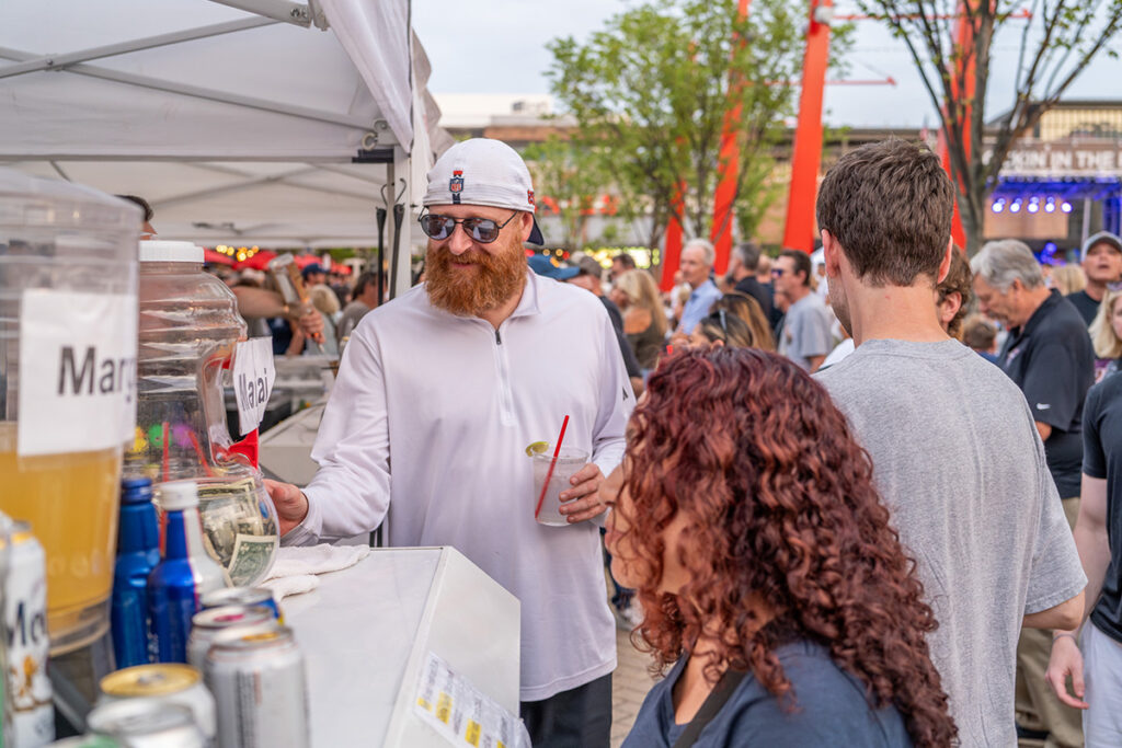 A man with a red beard and sunglasses buys a drink at an outdoor festival booth, surrounded by a crowd. Various beverages and jars are visible on the counter, and trees and stage lights appear in the background.
