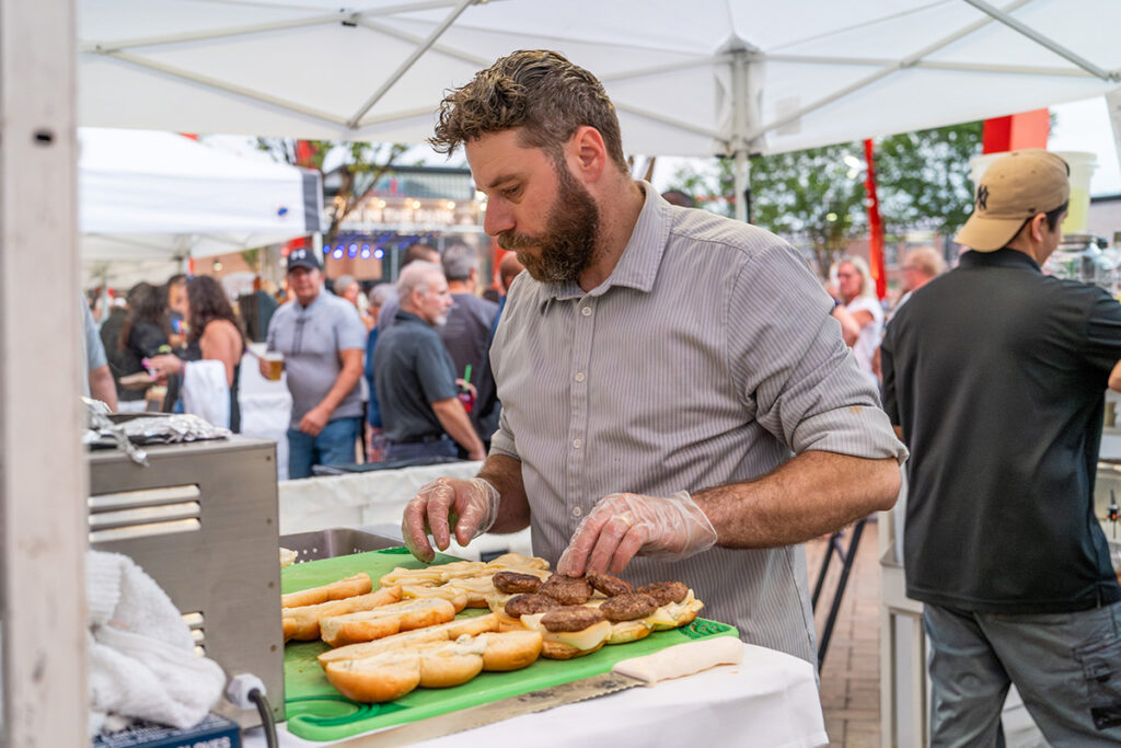 A man wearing gloves prepares sandwiches at an outdoor event, slicing bread and placing meat on buns at a table under a white tent, while a crowd gathers in the background.