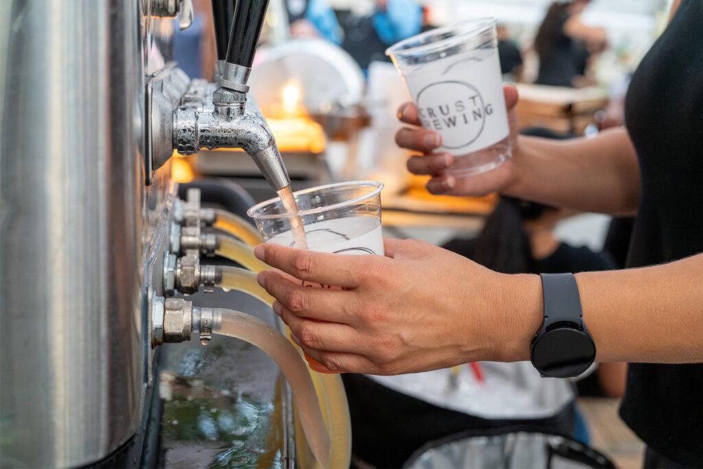 A person fills a plastic cup with beer from a tap at a brewery event. Another cup with a brewery logo is held in their other hand. The person wears a black smartwatch. Other people are blurred in the background.