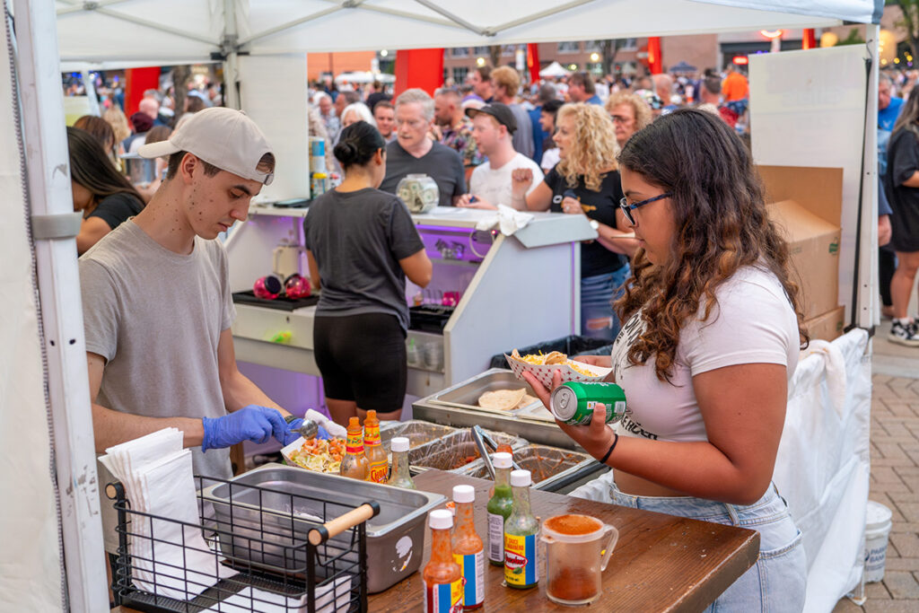 A young man prepares food at a busy outdoor food stall while a woman holding a can waits, surrounded by bottles of hot sauce. A crowd of people gathers in the background under white tents.
