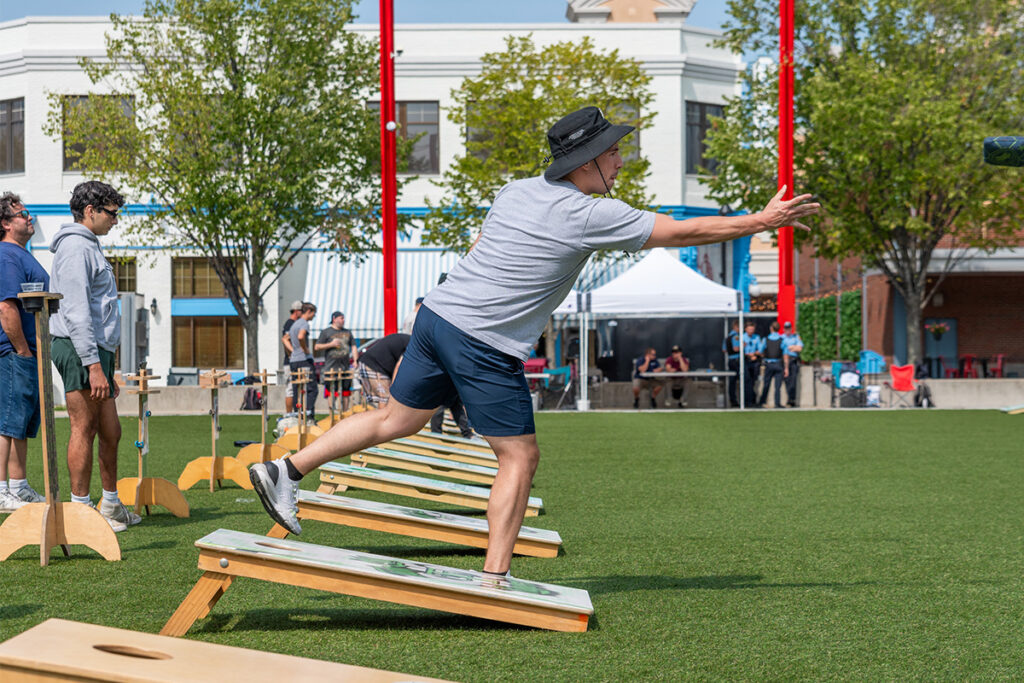 A man wearing a hat, gray shirt, and shorts throws a beanbag while playing cornhole on a grassy outdoor field. Several people watch in the background near buildings and trees on a sunny day.