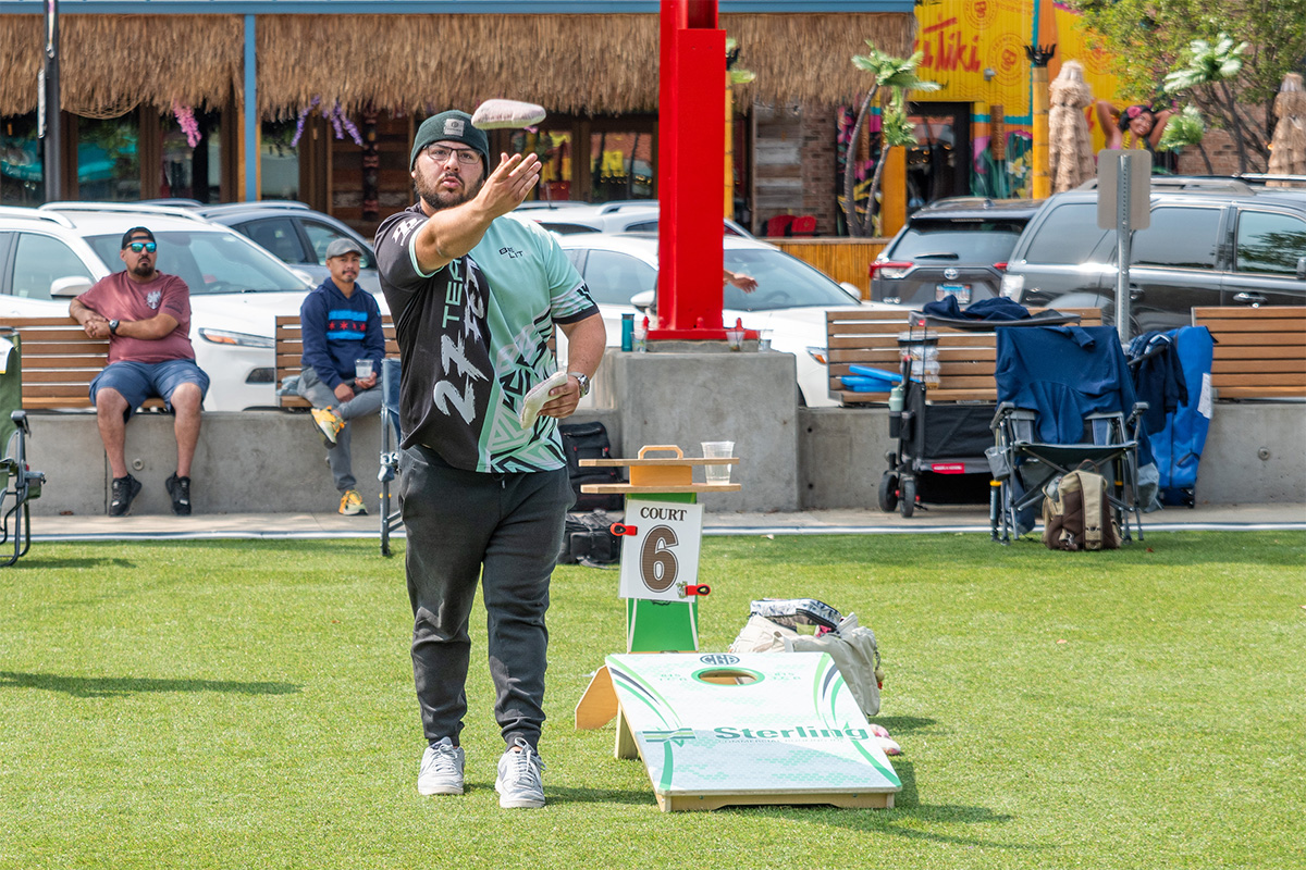 A man wearing a green and black jersey tosses a bean bag toward a cornhole board on a grassy court labeled “Court 6,” with spectators and parked cars in the background.