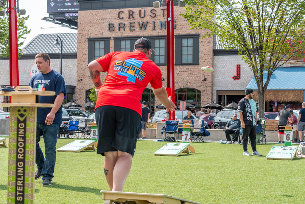 People play cornhole on a grassy area outside a brewery, with one person in a red shirt throwing a beanbag. Others watch or wait their turn. Folding chairs and parked cars are visible in the background.
