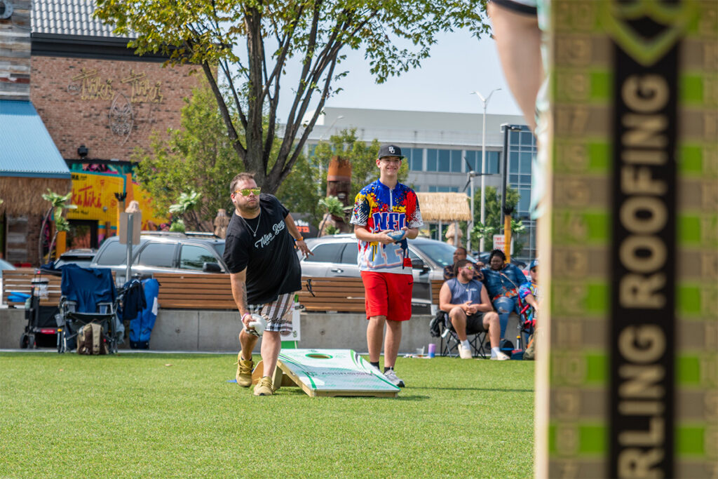 Two men play cornhole on a grassy field; one is tossing a bean bag while the other stands nearby. People sit in chairs in the background, and there are buildings, cars, and trees surrounding the scene.