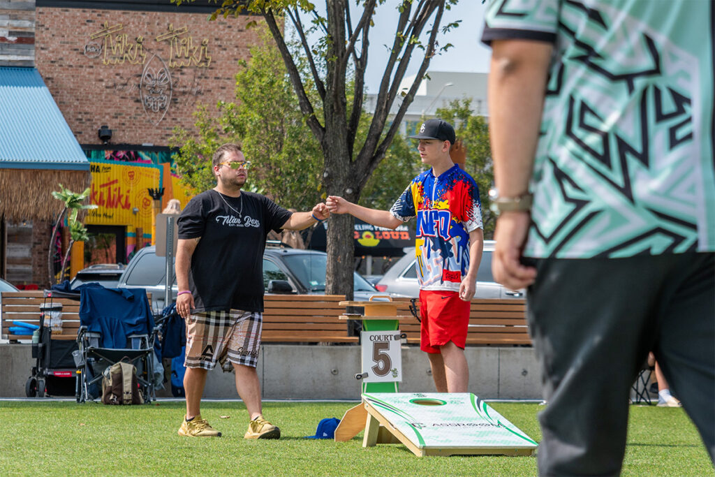 Two men standing on grass fist bump near a cornhole board, one wearing a black shirt and shorts, the other in a colorful sports jersey and red shorts. Another person in a green patterned shirt is partially visible in the foreground.