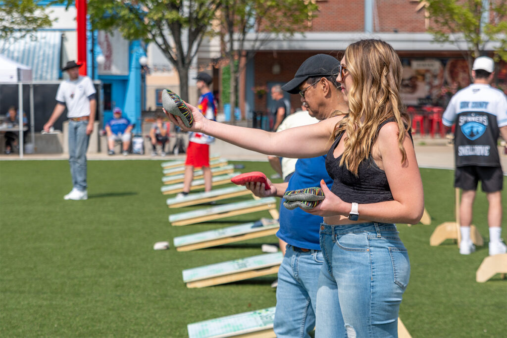 Two people stand side by side outdoors playing cornhole, each holding and tossing a bean bag toward wooden boards. Other people and cornhole boards are visible in the background on a grassy area.