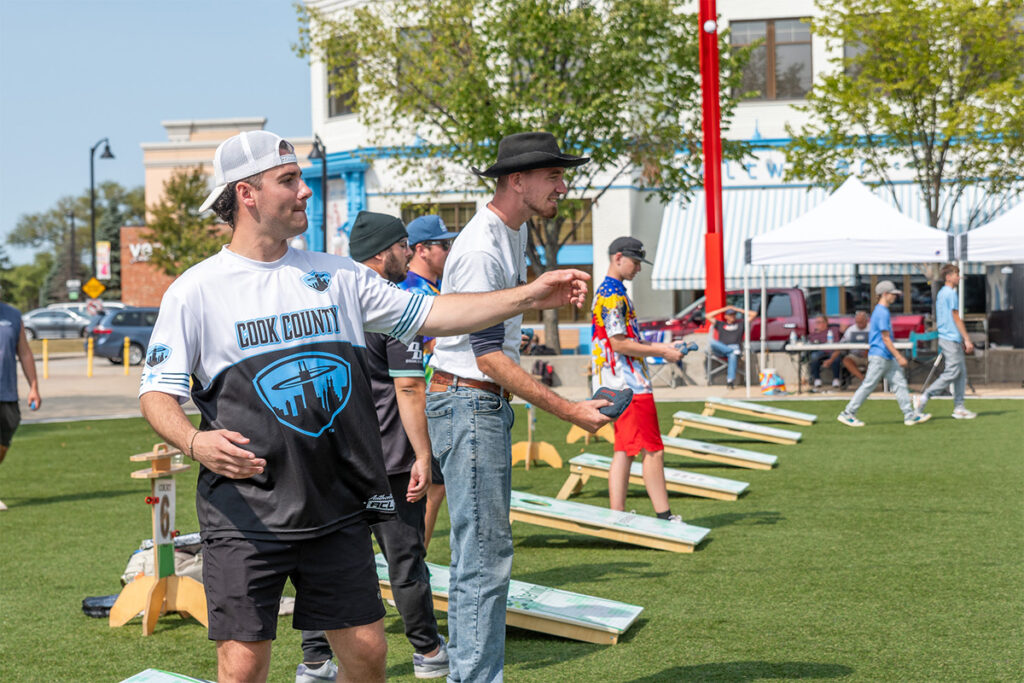 Several people play cornhole outdoors on a sunny day. The foreground shows a man in a Cook County jersey tossing a beanbag, while others wait their turn. There are buildings and trees in the background.