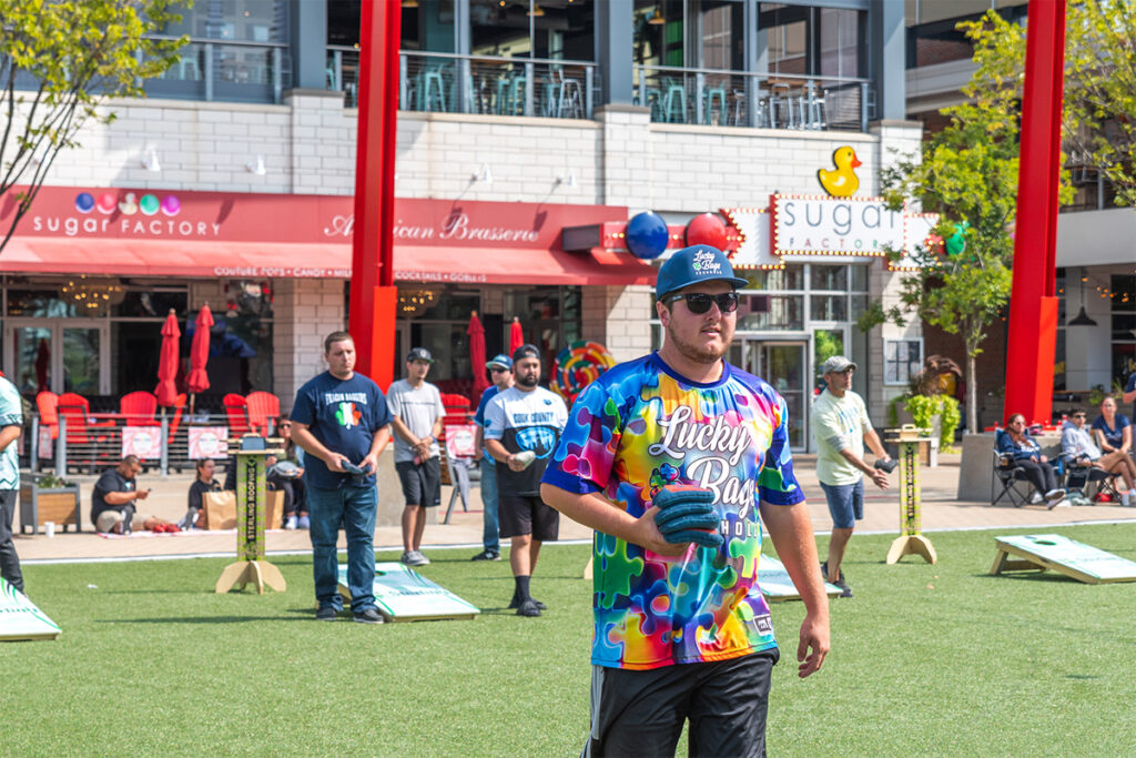 A man wearing sunglasses and a colorful Lucky Bags shirt holds a bean bag on a grassy area, while others play cornhole in the background near a restaurant with red umbrellas.