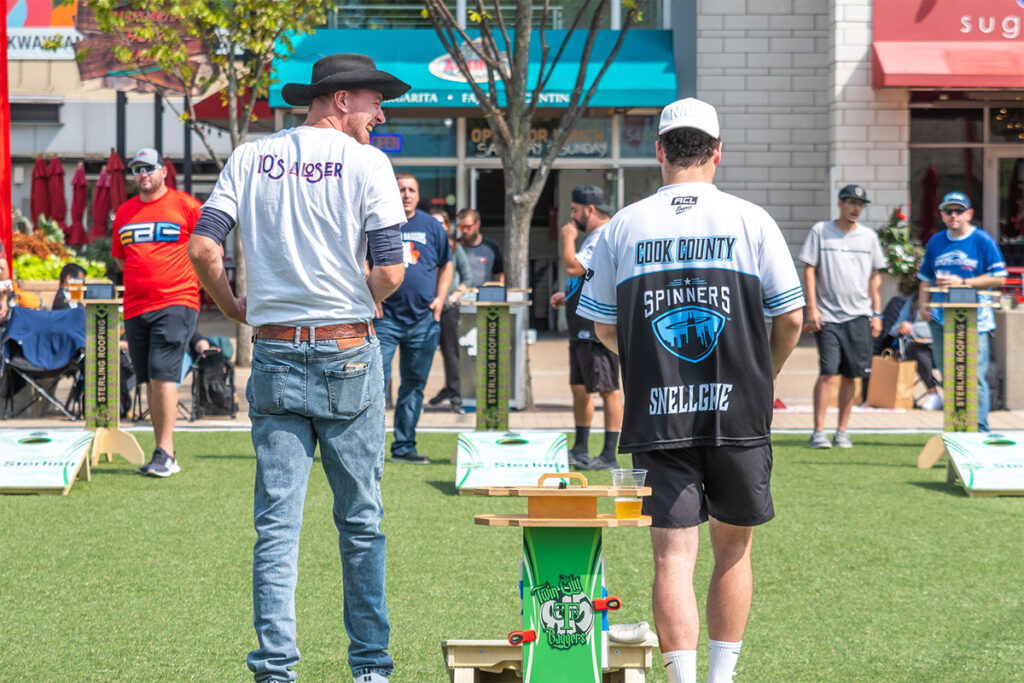 Two men stand on artificial grass at an outdoor event, one in jeans and a cowboy hat, the other in a sports jersey. People and games are visible in the background, with a sunny and lively atmosphere.