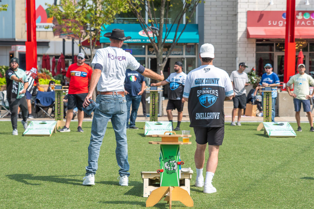 Two men play cornhole on a grassy field as a crowd watches. One man tosses a bean bag while the other stands nearby. Several cornhole boards and onlookers are visible in the background on a sunny day.