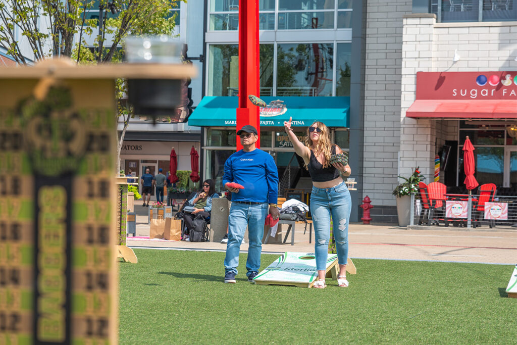 A woman throws a bean bag while playing cornhole with a man outdoors on a lawn, with shops and people in the background on a sunny day.