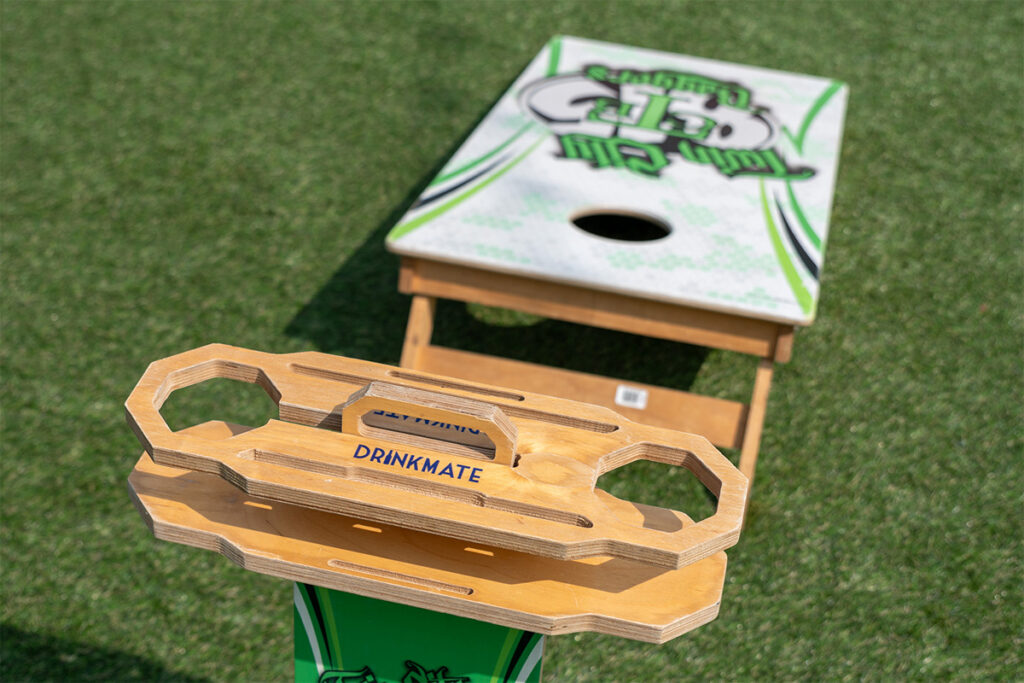A wooden drink holder labeled DRINKMATE is positioned on a grassy field, with a cornhole board featuring a green and white design visible in the background.