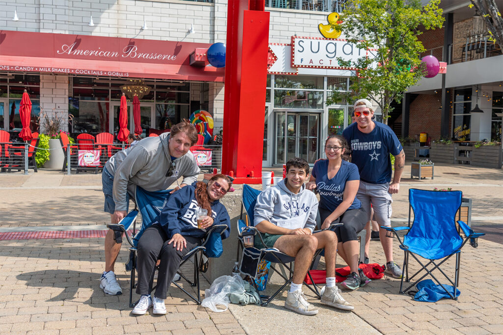 Six people sit and smile for a group photo on folding chairs outdoors in front of restaurants, with a red sculpture and balloons in the background. It is a sunny day, and some wear Dallas sports shirts.