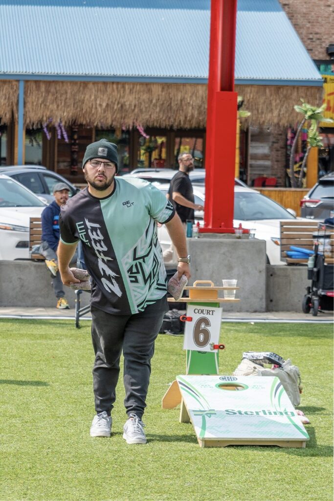 A man wearing glasses and a green-and-black shirt tosses a beanbag while playing cornhole on a grassy field. A “Court 6” sign is next to the board, and parked cars and a building are in the background.