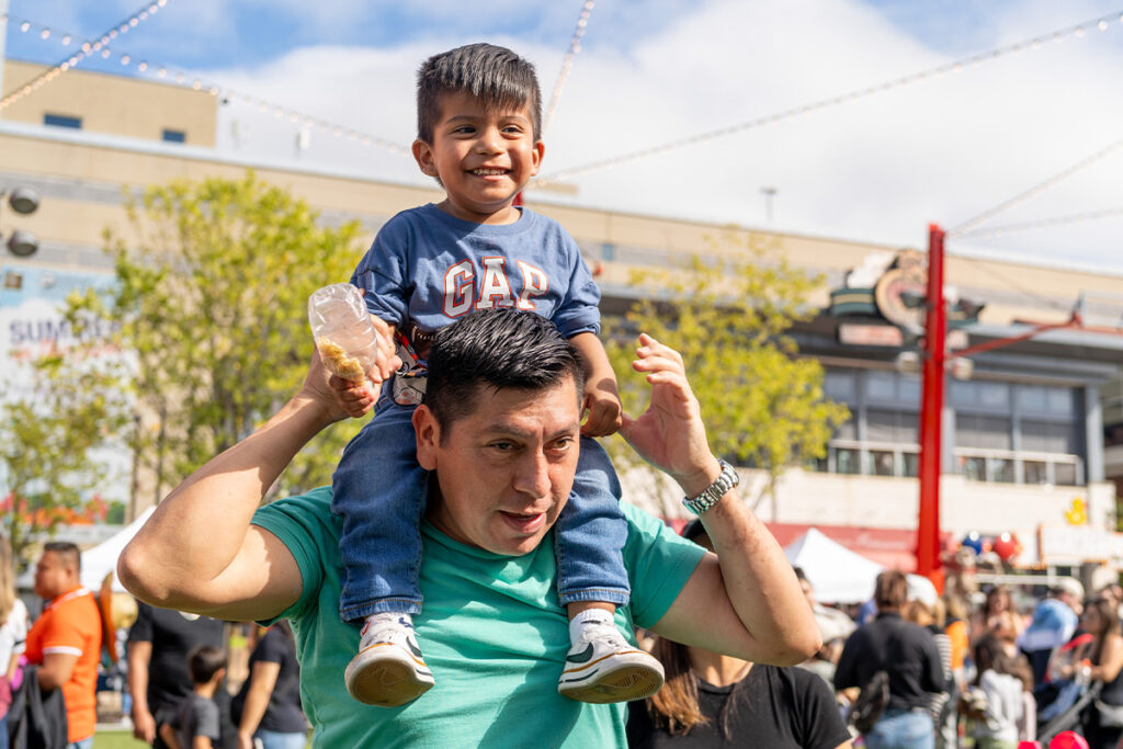 A smiling man in a green shirt carries a happy young boy on his shoulders at an outdoor festival. The boy holds a bag of snacks, and people and tents are visible in the background.
