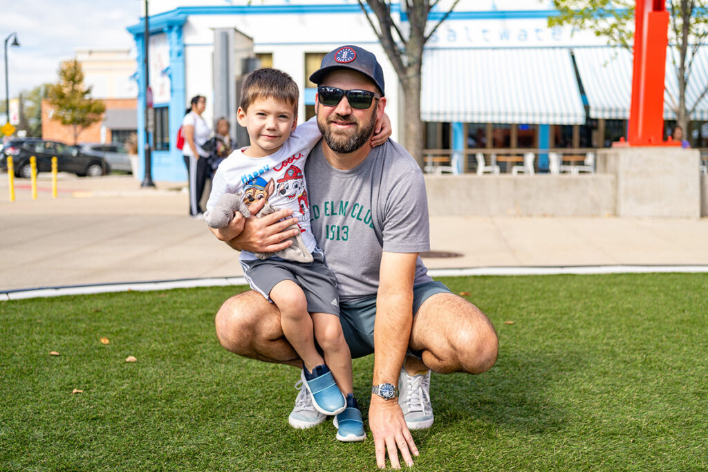 A man in sunglasses and a gray cap kneels on grass, hugging a young boy who is smiling and holding a stuffed toy. They are outdoors, with shops and trees in the background on a sunny day.