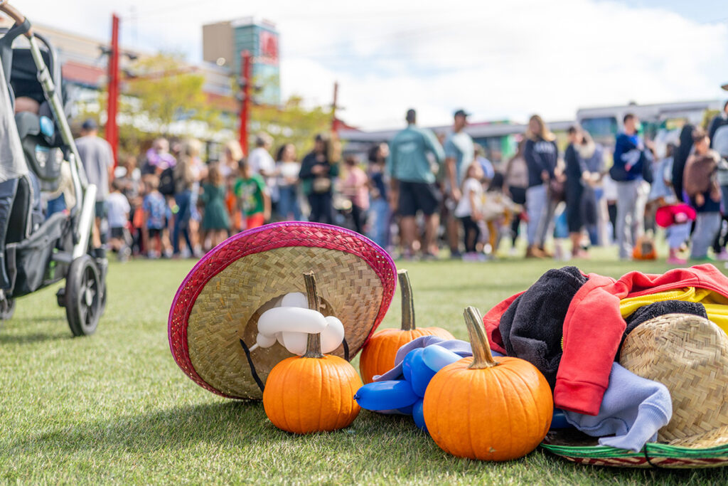 A pile of small pumpkins, hats, balloon animals, and colorful fabric lays on grass in the foreground, with a crowd of people and children gathered outdoors in the background.