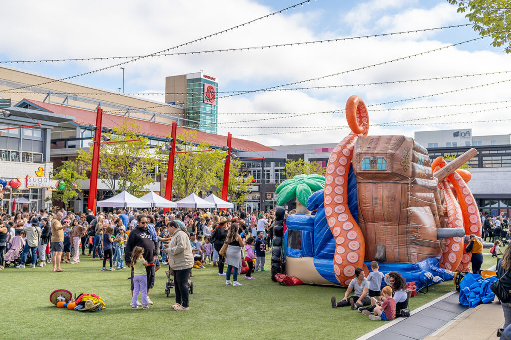 A crowd of people gathers in an outdoor plaza with white tents, string lights, and a large inflatable pirate ship featuring octopus tentacles. Children and families enjoy activities on the grass under a partly cloudy sky.
